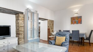 Un salon avec une table basse en verre, un canapé bleu, un coin repas avec une table et des chaises, un téléviseur et un meuble.
