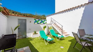 Une terrasse avec un revêtement de gazon synthétique, deux fauteuils de jardin verts, un barbecue et un escalier menant à un bâtiment blanc dont la façade est recouverte de carreaux bleus et verts.