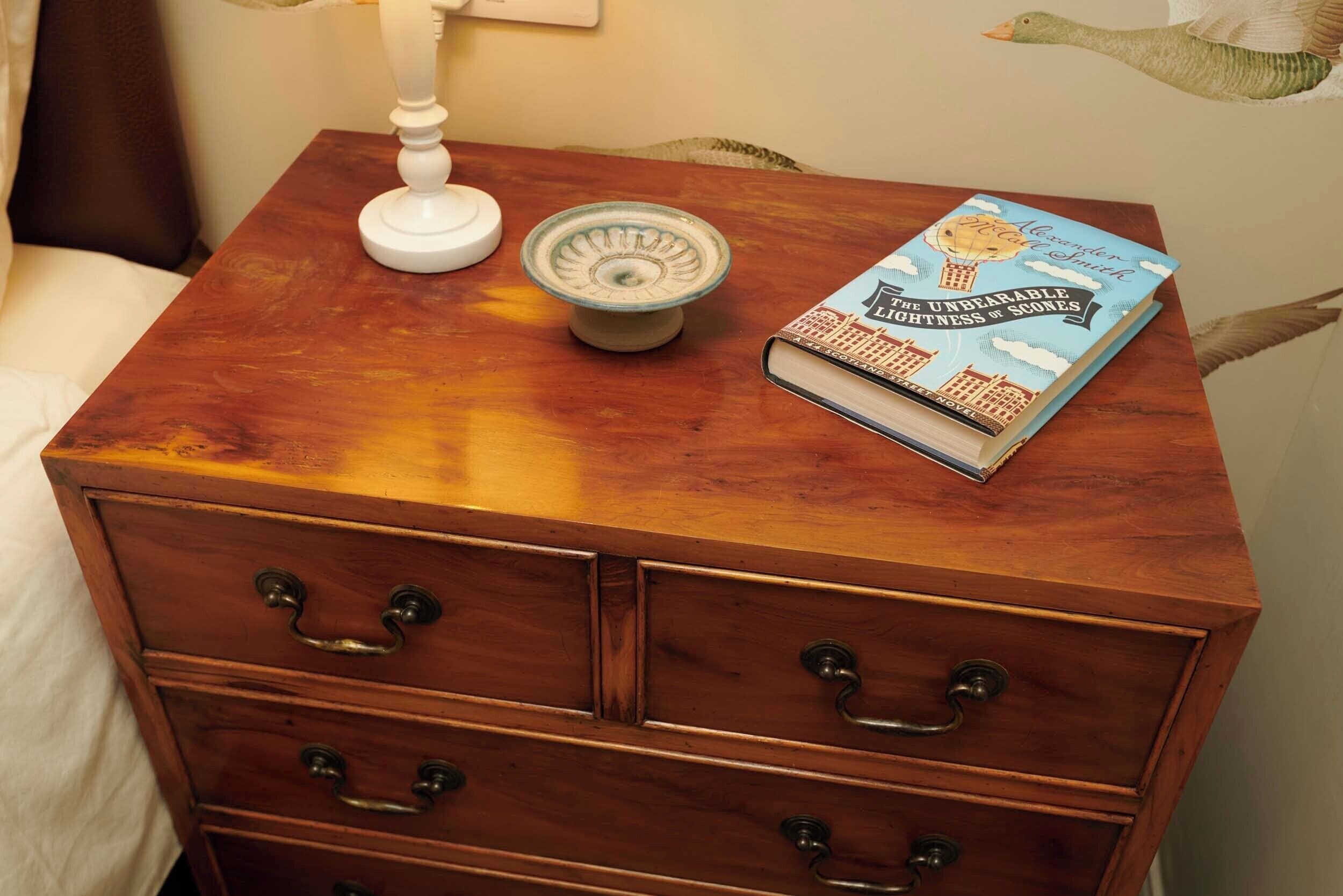 Een houten nachtkastje met een boek getiteld 'The Unbearable Lightness of Scones' en een witte lamp.