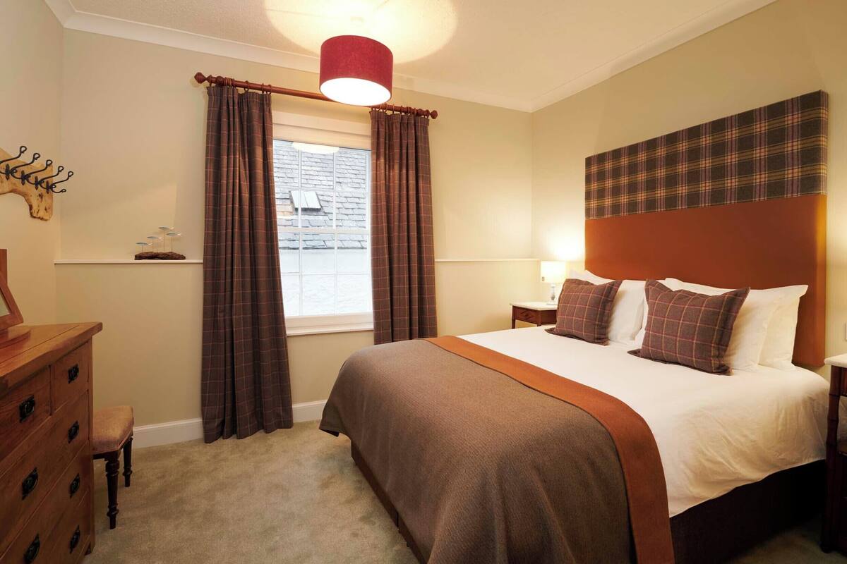 A neatly arranged bedroom with a large bed, a wooden dresser, a nightstand, and a window with curtains.