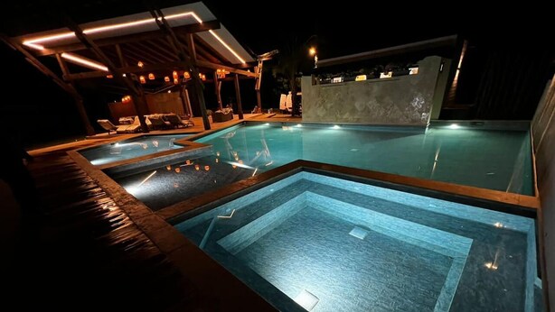 A swimming pool area with a wooden deck and lounge chairs, surrounded by a covered structure with lighting.