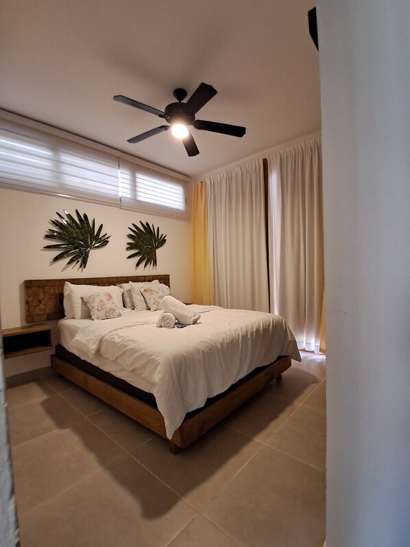 Una cama bien hecha con sábanas blancas y un cabecero de madera, un ventilador de techo y dos plantas decorativas colgadas en la pared.