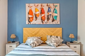 A hotel room with a bed, two nightstands, a wooden headboard, a blue wall, and a painting of fish. - Apartment 'Sea View Apartment - Beach 30m Away' with Ocean View, Private Terrace & Wi-Fi (Cléder)