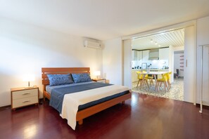 A bedroom with a wooden bed, bedside tables, and a view into a dining area with yellow chairs.