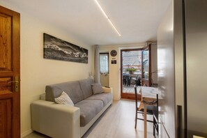 Un salon moderne avec un canapé, une table à manger et des chaises, une horloge, et une vue sur un espace extérieur accessible par une porte vitrée.