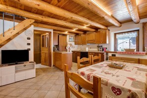 Une cuisine avec des meubles de rangement en bois, une table à manger recouverte d’une nappe à motifs et un téléviseur posé sur un meuble.