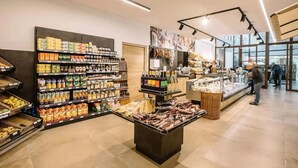 A grocery store interior with shelves stocked with various products, a central display case, and a visible entrance.