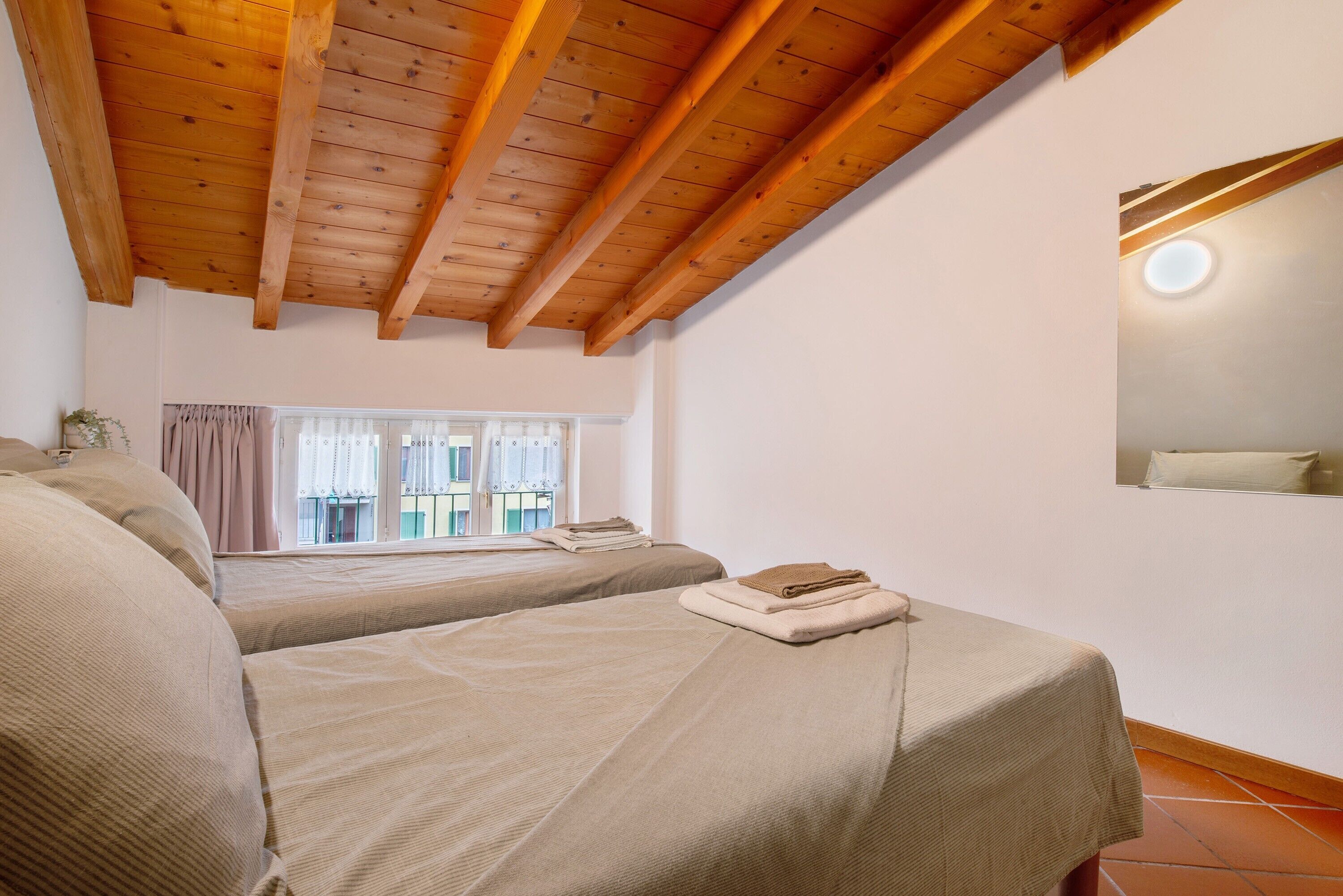 Ein Schlafzimmer mit zwei Betten, Holzbalkendecke, einem Fenster mit Vorhängen und einer Wandleuchte.