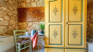 A stone wall, a wooden cabinet with floral designs, a green chair, a white table, and a kettle.
