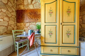 A stone wall, a wooden cabinet with floral designs, a green chair, a white table, and a kettle. - Room 'Masseria Copertini - Angelo' with Wi-Fi and Air Conditioning (Vernole)