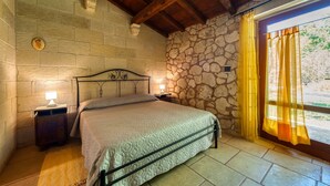 A bedroom with a stone wall, a wooden ceiling, a bed with a metal frame, two bedside tables with lamps, and a window with curtains.