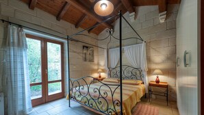 Un dormitorio con cama con dosel, techo de madera, paredes de piedra, una ventana con cortinas y una mesita de noche con lámpara.