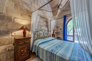 Un dormitorio con pared de piedra, una cama con colcha azul y blanca a rayas, una mesita de noche con lámpara y una ventana con cortinas azules.