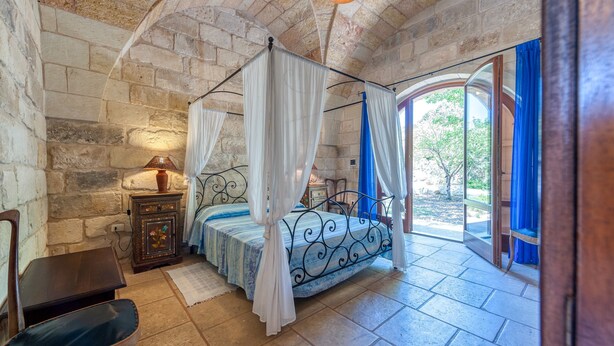 A bedroom with a stone wall, a bed with a canopy, a nightstand, a chair, and a view through a glass door. - Room 'Masseria Copertini - Gattini' with Wi-Fi and Air Conditioning (Vernole)