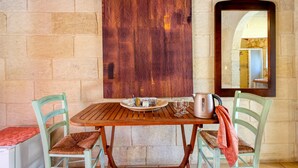 Een houten tafel met een bord, glazen en een waterkoker, omringd door twee stoelen met kussens, tegen een stenen muur met een groot abstract schilderij en een spiegel.