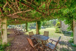 Un espace extérieur abrité, avec une table et des chaises, entouré de verdure et d’un mur en pierre.