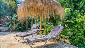 Een terras met twee loungestoelen, een rieten parasol en weelderige begroeiing.