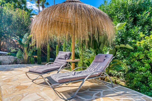 A patio with two lounge chairs, a thatched umbrella, and lush greenery.