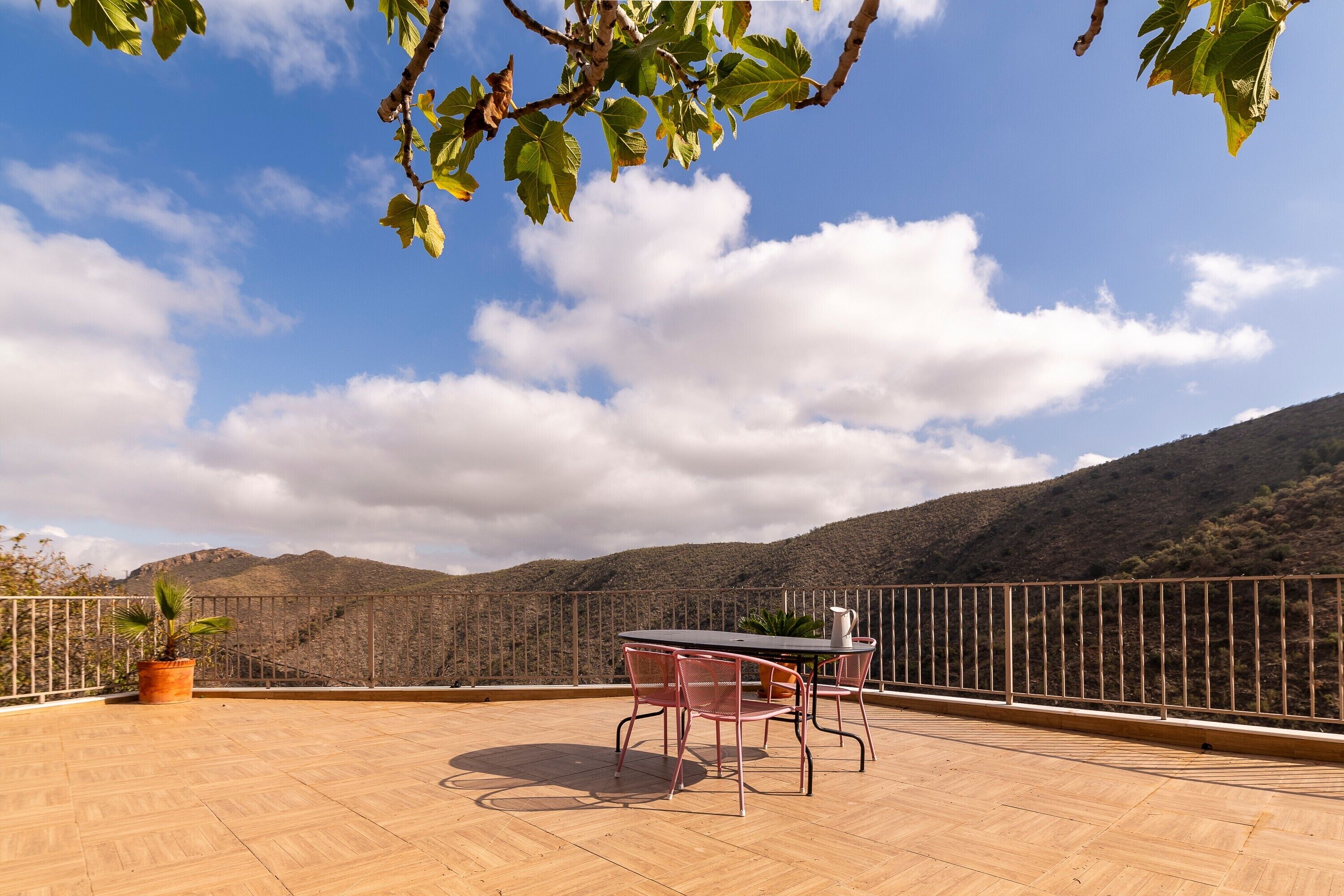 A terrace with a table and chairs, a potted plant, and a view of a mountainous landscape.