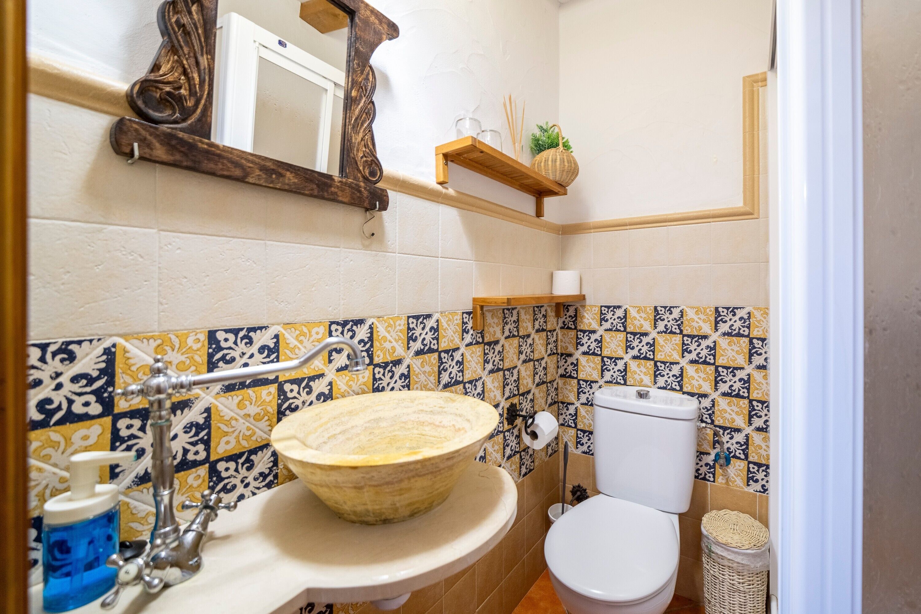 A bathroom with a ceramic sink, a toilet, a mirror, and a shelf with a plant.