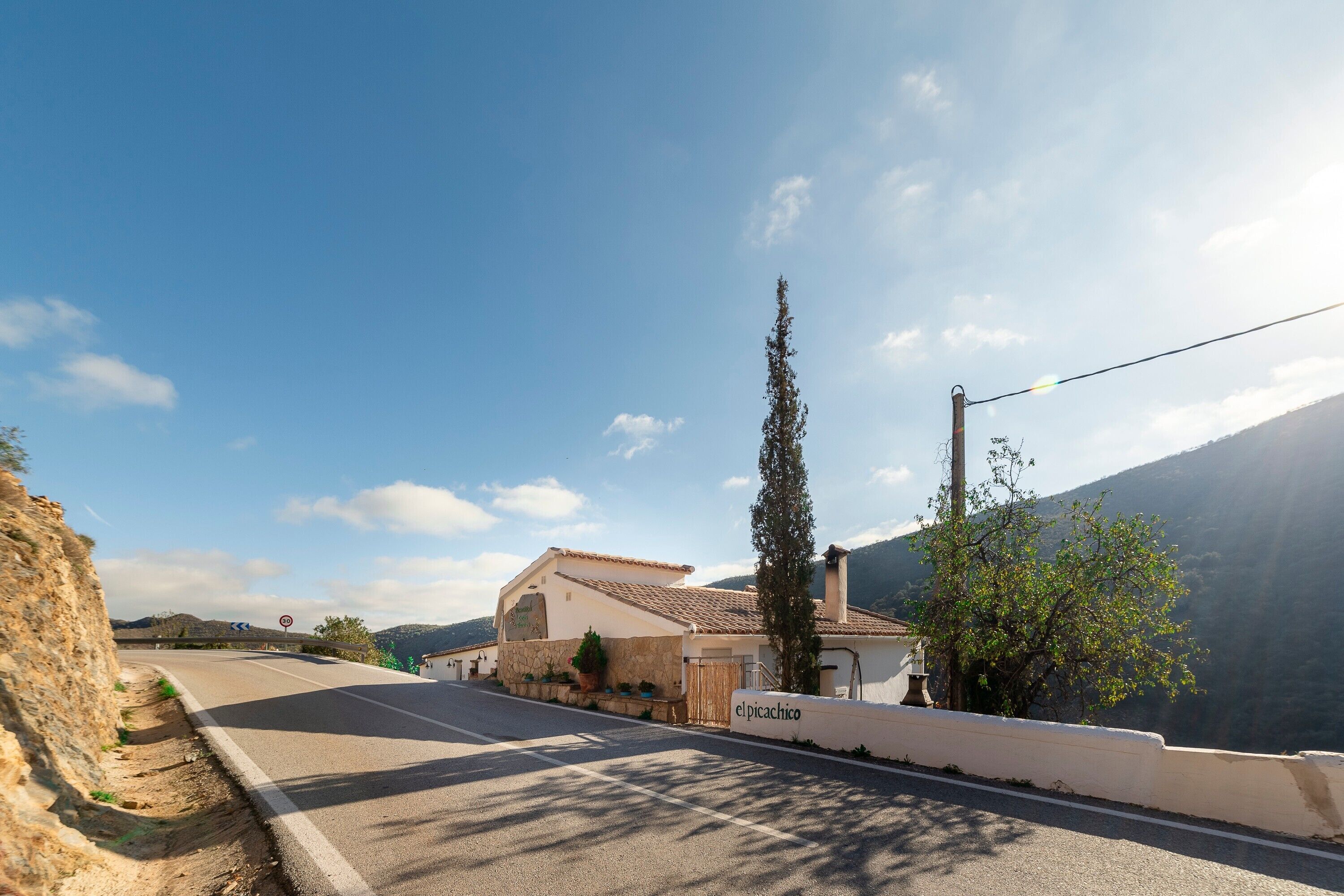 Un camino rural con un edificio blanco identificado como 'el picachico', un muro de piedra y un árbol.