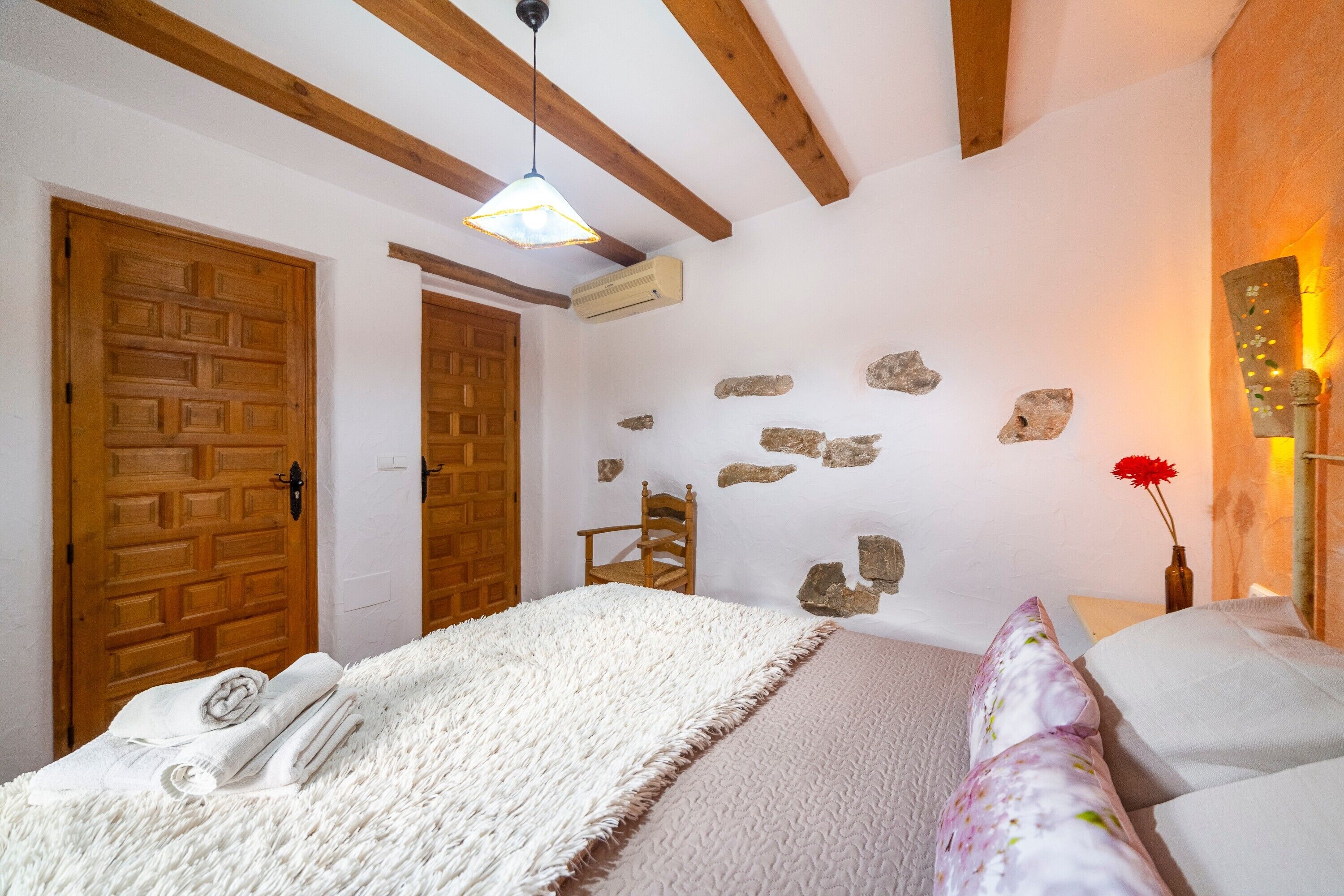 Un dormitorio con puerta de madera, cama con manta blanca, pared de piedra y lámpara colgante.