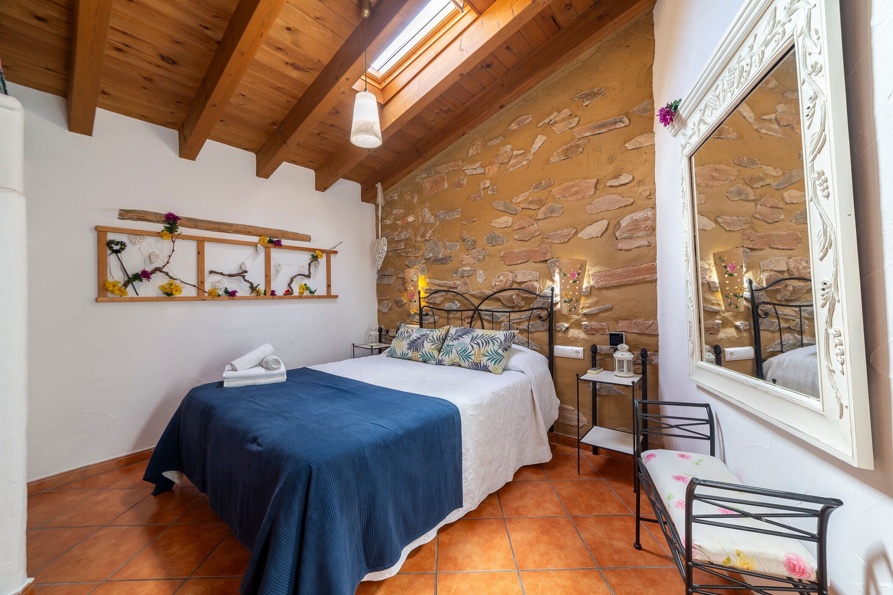 Une chambre à coucher comprenant un lit, une table de chevet, un miroir, une étagère murale agrémentée d’objets décoratifs et un mur en pierre.
