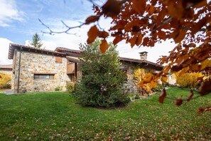 Un bâtiment en pierre avec une porte et une fenêtre en bois, entouré d’une cour verdoyante et d’arbres.
