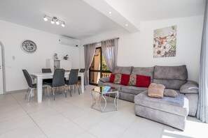 A modern living room with a grey sofa, a coffee table, a dining area with chairs, and a wall clock.