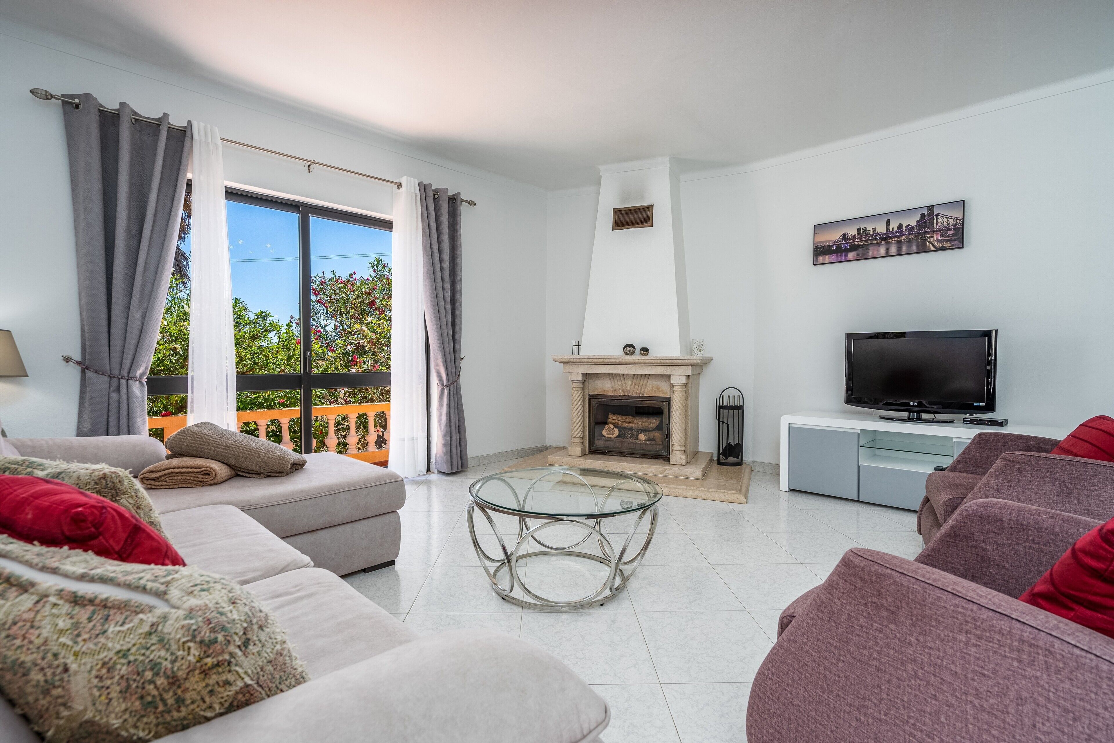 A living room with a sofa, armchair, glass coffee table, fireplace, TV, and a balcony with curtains.