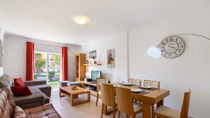 A living room with a sofa, a dining table with chairs, a clock on the wall, and a TV.