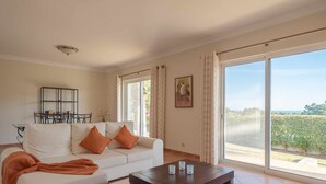 A living room with a white sofa, orange cushions, a coffee table, a bookshelf, and large windows with a view of greenery.