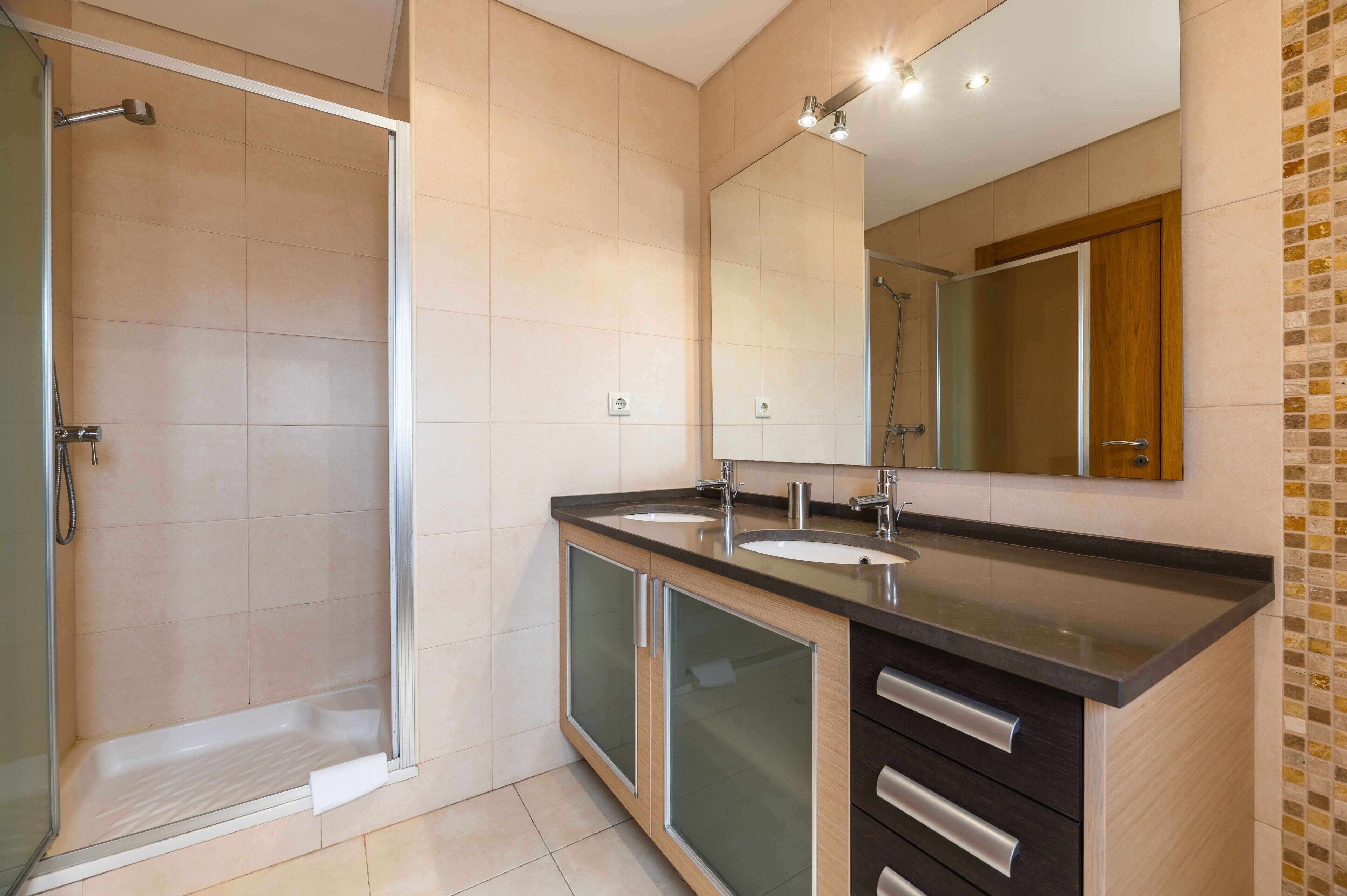 Een moderne badkamer met een douche, twee wastafels, een grote spiegel en een kast met lades.