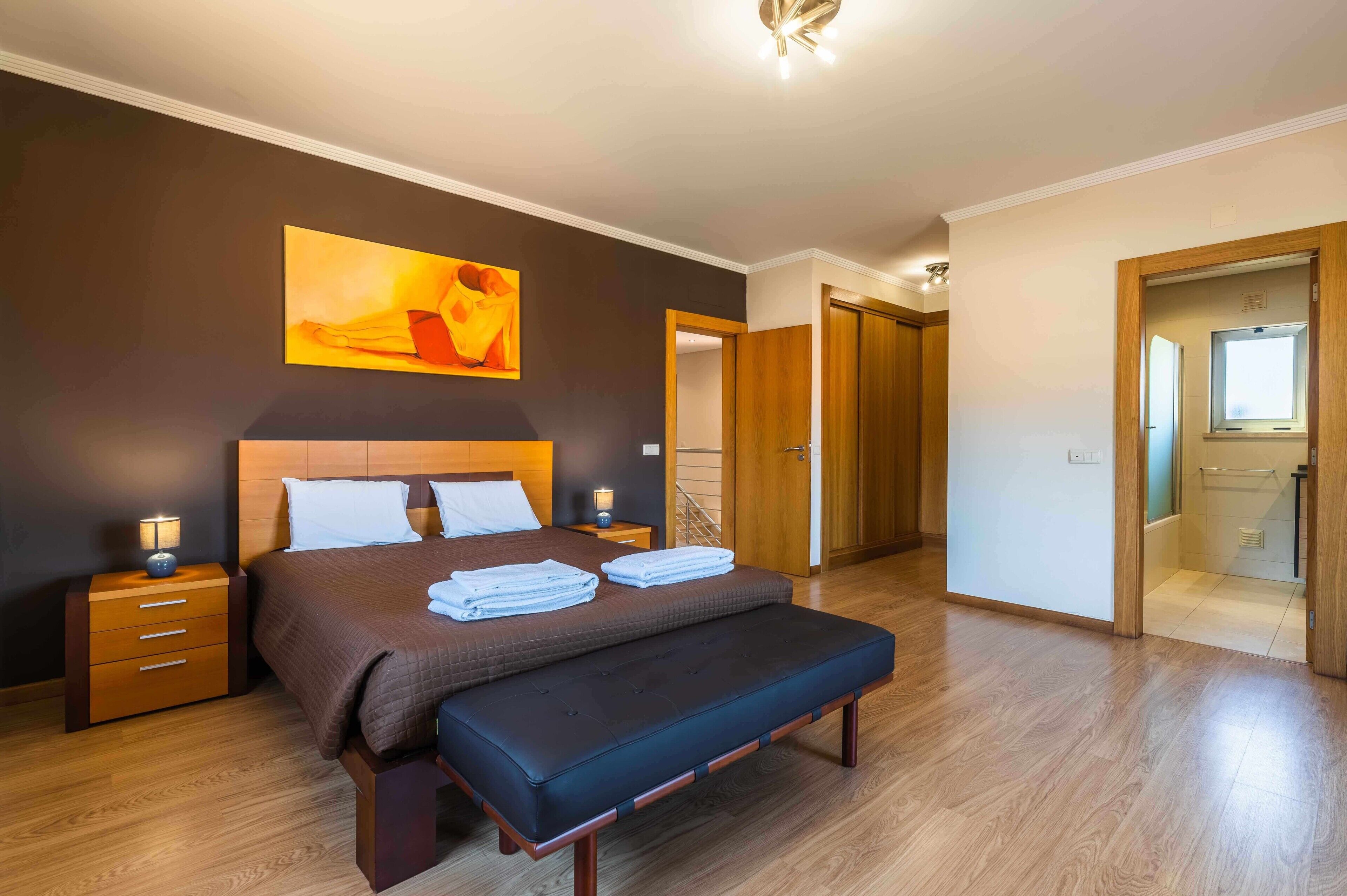 Un dormitorio con cama, mesitas de noche, un banco, un cuadro y una puerta que da a un baño.
