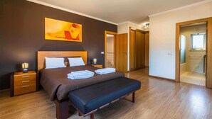 Un dormitorio con cama, mesitas de noche, un banco, un cuadro y una puerta que da a un baño.