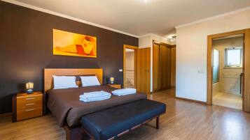 Un dormitorio con cama, mesitas de noche, un banco, un cuadro y una puerta que da a un baño.