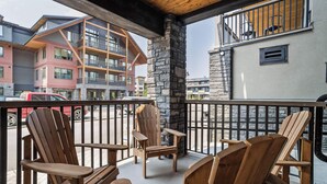 A balcony with wooden Adirondack chairs, a stone pillar, and a view of multi-story buildings. - 114 Two Bedroom Premier Condo (Canmore)