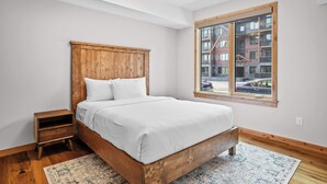 A bedroom with a wooden bed, a nightstand, a window with a view of buildings, and a ceiling light. - 114 Two Bedroom Premier Condo (Canmore)