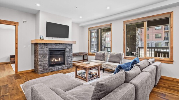 A modern living room with a stone fireplace, a large flat-screen TV, and a large window offering a view of buildings. - 114 Two Bedroom Premier Condo (Canmore)