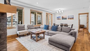 A spacious living room with a large sectional sofa, a coffee table, and a fireplace. There are large windows with wooden frames, a dining area, and a door leading to another room. - 114 Two Bedroom Premier Condo (Canmore)