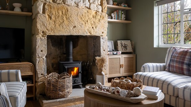 A cozy living room with a stone fireplace, wicker baskets, a striped sofa, and a window with a view of greenery. - Broadway Scandi Chic Cottage (Broadway)