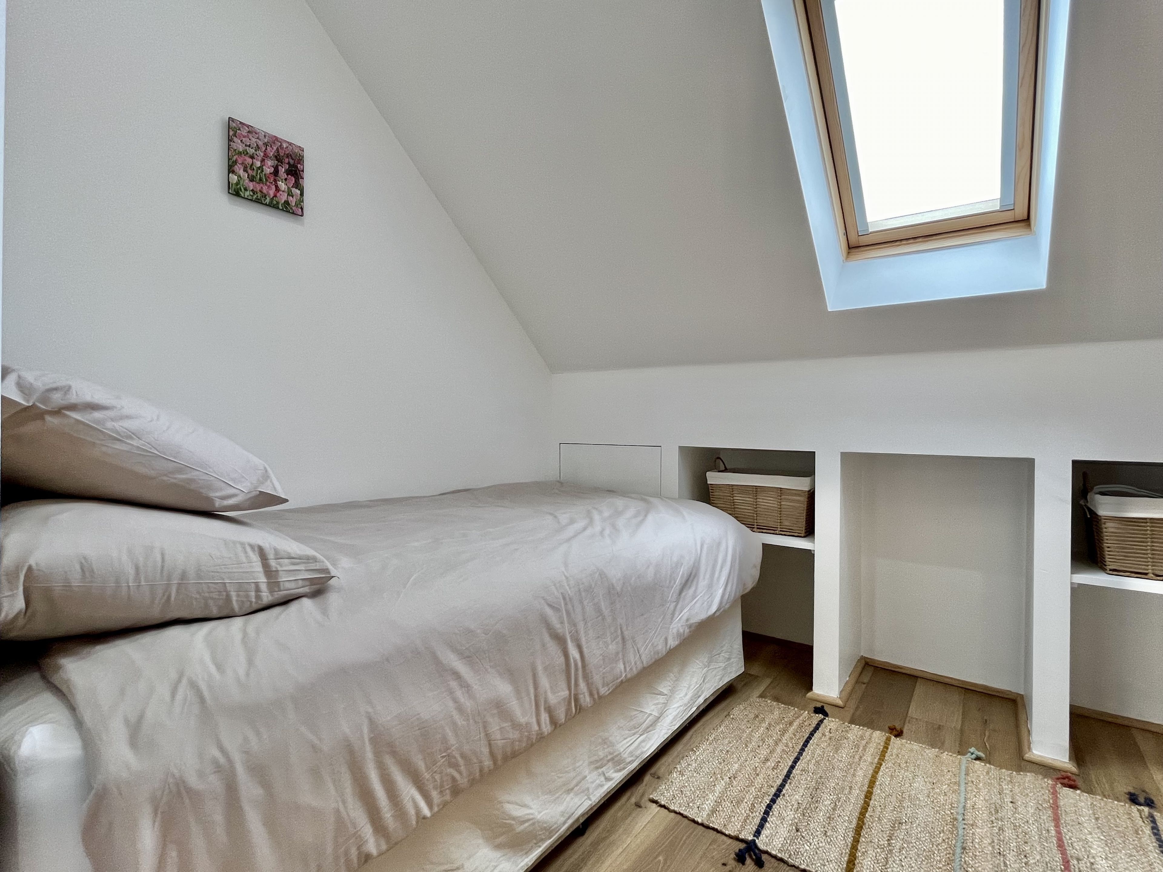 A bedroom with a sloped ceiling, a bed with white bedding, a small picture on the wall, and built-in storage shelves.