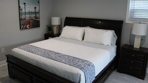 Une chambre avec un grand lit, deux tables de chevet, une petite table d’appoint, une lampe fixée au mur et un tableau représentant une scène de plage.