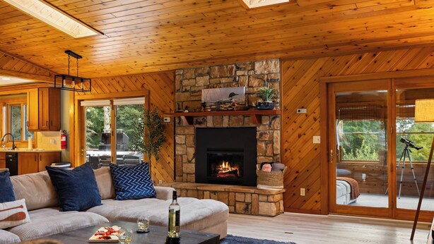L’intérieur chaleureux d’un chalet avec une cheminée en pierre, un plafond en bois et une grande fenêtre donnant sur une terrasse.