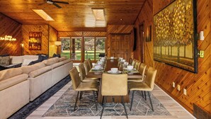 Un coin repas avec une longue table dressée pour six personnes, entourée de six chaises, dans une pièce aux murs et au plafond en bois, dotée d’un ventilateur de plafond et d’une grande fenêtre offrant une vue sur la verdure environnante.