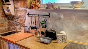 Cozinha com parede de pedra, tábua de corte de madeira, utensílios pendurados, máquina de café, torradeira e janela com vista.