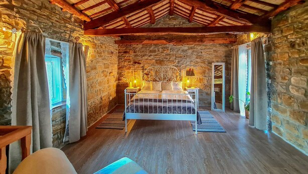 Quarto com paredes de pedra, teto de madeira, cama, abajures e vista para uma parede de pedra.
