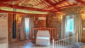 Quarto rústico com parede de pedra, teto de madeira, cama, uma mesinha com uma planta e uma janela com cortinas.
