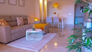 Ein modernes Wohnzimmer mit einer Couch, einem Couchtisch und Sesseln. Im Hintergrund befindet sich ein Essbereich mit Tisch und Stühlen.