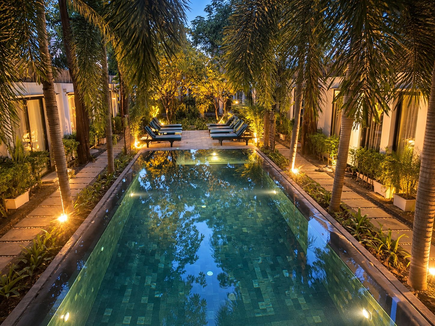A rectangular swimming pool with a tiled edge, surrounded by palm trees and benches.